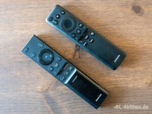 Oben die Fernbedienung des Samsung S90H OLED TV mit AI-Assistent-Taste und Solarpanel auf der Rückseite.